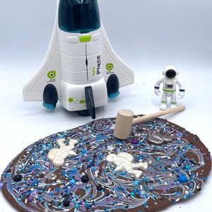 May include: A white toy spaceship with a green logo that says "Plane" is on a white surface next to a chocolate space-themed bark with white chocolate astronaut figures and sprinkles. A wooden mallet is on the bark and a white toy astronaut is standing on the surface.
