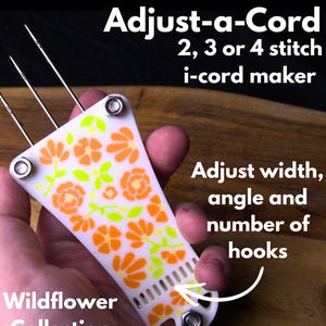 May include: A white i-cord maker with orange and green floral patterns, held in a hand. The text "Adjust-a-Cord" and "Wildflower Collection" are visible. It is designed for 2, 3, or 4 stitch i-cord projects. The tool allows adjustment of width, angle, and number of hooks.