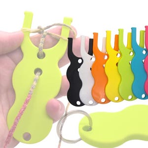 Puede incluir: Un conjunto de herramientas de plástico de colores para hacer pulseras de la amistad. Las herramientas tienen forma de ocho y tienen dos agujeros. Las herramientas se muestran en una variedad de colores, incluyendo negro, blanco, naranja, amarillo, verde, azul, rosa, morado y gris.