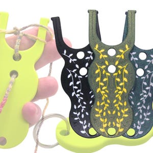 May include: Three different colored plastic tools for making friendship bracelets. The tools are shaped like a U with a hole in the middle. The tools are decorated with a leafy pattern. The tools are on a yellow plastic base.