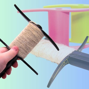 May include: A black yarn swift with a spool of beige yarn is held in a hand. The swift has two black arms that extend from the center. In the background are two colorful yarn spools, one pink and one green.