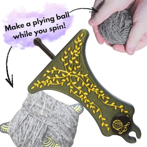 Puede incluir: Una herramienta para torcer hilo verde y dorado con un mango marrón oscuro y una madeja de hilo gris. La herramienta tiene un diseño de hojas decorativas y el texto "Make a plying ball while you spin!"