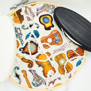 May include: A wooden embroidery hoop with a white fabric background featuring a colorful pattern of musical instruments, including guitars, violins, and a trumpet. A white and a black oval-shaped piece of fabric are attached to the hoop.