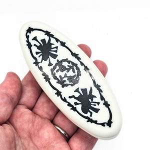 May include: White oval-shaped trinket box with a black and white design of stylized flowers and leaves. The design is framed by a black barbed wire border.