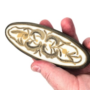 Puede incluir: Un objeto decorativo de madera de forma ovalada con fondo blanco y diseño floral negro y dorado.