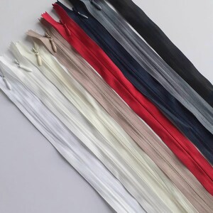 May include: Assortment of zipper closures in various colors, including white, beige, cream, red, navy blue, gray, and black. The zippers are laid out in a diagonal line on a white background.
