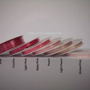 May include: A collection of eight spools of ribbon in various shades of pink, fuchsia, and peach. The spools are arranged in a row, with the darkest shade of pink on the left and the lightest shade of pink on the right. The spools are labeled with their respective colors: Pink, Fuschia, Light Pink, Baby Pink, Peach, Light Peach, and Hinted Pink.
