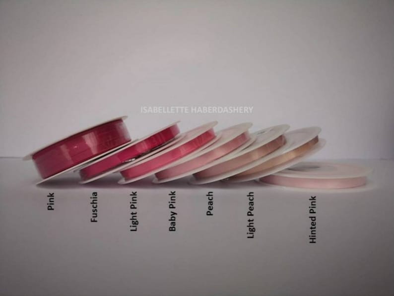May include: A collection of eight spools of ribbon in various shades of pink, fuchsia, and peach. The spools are arranged in a row, with the darkest shade of pink on the left and the lightest shade of pink on the right. The spools are labeled with their respective colors: Pink, Fuschia, Light Pink, Baby Pink, Peach, Light Peach, and Hinted Pink.