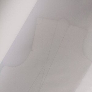 May include: A light-colored sewing pattern, possibly for a bodice or top, is visible. The pattern is drawn on a translucent material, with visible lines and shapes indicating the design. The overall image is softly lit, with a focus on the pattern's details.