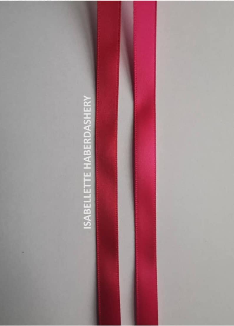 May include: Two satin ribbons, one in a deep pink and the other in a slightly lighter shade of pink. The ribbons are both about 1 cm wide and are lying flat on a white surface. The text "ISABELLETTE HABERDASHERY" is printed vertically on the left side of the image.