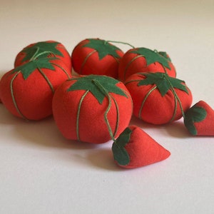 May include: A group of seven red fabric pincushions shaped like tomatoes and strawberries. The pincushions have green felt leaves and are sewn with white thread.