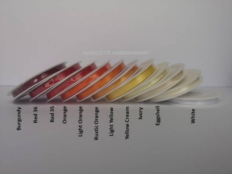 May include: A collection of spools of ribbon in various colors, including burgundy, red, orange, yellow, cream, ivory, eggshell, and white. The spools are arranged in a row from darkest to lightest.
