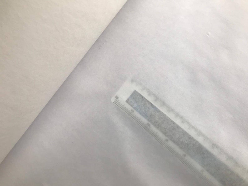 May include: A close-up shot of white fabric with a ruler placed diagonally across the material. The ruler is gray with black markings, indicating measurements in inches and centimeters. The fabric appears soft and slightly textured.