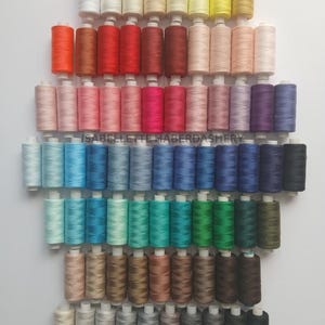 May include: A collection of 100 spools of thread in various colours, arranged in rows. The spools are white with a clear plastic window showing the thread colour. The thread colours range from white to black, with shades of red, pink, blue, green, brown, and grey. The text "ISABELLETTE HABERDASHERY" is visible in the centre of the image.