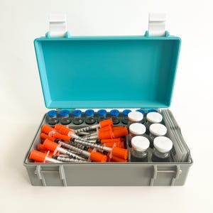 May include: An open, teal and gray medical kit containing vials with blue caps, syringes with orange caps, and vials with white caps. The kit is on a white background.