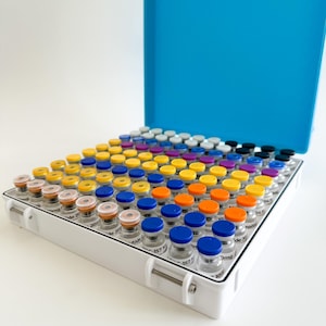 May include: A white case with a blue lid, filled with numerous small glass vials. The vials have colorful caps in shades of yellow, orange, blue, purple, and white. The case is open, revealing the contents inside.