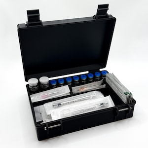 May include: A black rectangular hard-shell carrying case with a hinged lid, containing multiple small glass vials with blue and white caps, several individually wrapped syringes, and a stack of small white paper packets organized in compartments.