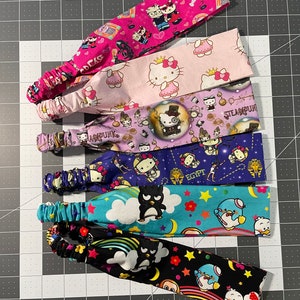 May include: Six colorful headbands with different cartoon characters and designs. The headbands are made of fabric and have a stretchy elastic band. The designs include Hello Kitty, a penguin, a rainbow, and a steampunk design.