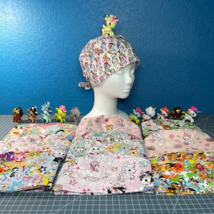 May include: A collection of colorful fabric squares with a variety of cartoon characters, including unicorns, cats, and other whimsical designs. The fabric squares are arranged in a row on a gray cutting mat. A small unicorn figurine is on top of a white surgical cap with a similar cartoon character print.