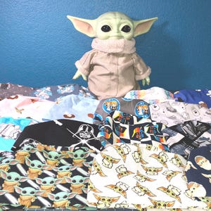 May include: A collection of 15 colorful fabric headbands featuring various Star Wars characters, including Yoda, Darth Vader, and Baby Yoda. The headbands are arranged in a pile with a plush Baby Yoda toy standing in front.