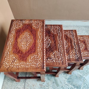 Könnte beinhalten: Ein Satz von vier verschachtelten Tischen aus Holz mit aufwendigen floralen Schnitzereien und eingelegten Perlmutteinlagen. Die Tische sind in absteigender Reihenfolge angeordnet, wobei der größte Tisch oben und der kleinste Tisch unten ist.