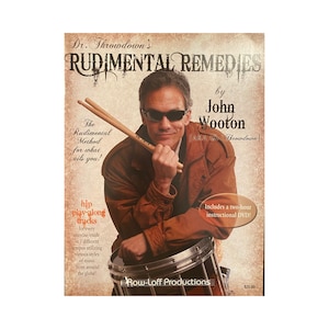 May include: A book cover with a brown background and a photo of a man holding drumsticks. The book title is "Rudimental Remedies" by John Wooton, AKA Dr. Throwdown. The book cover also includes the text "The Rudimental Method for what ails you!" and "Includes a two-hour instructional DVD!"