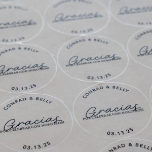 Peut inclure: Autocollants ronds transparents avec texte noir. Les autocollants indiquent "Conrad & Belly" et "Gracias Por Celebrar Con Nosotros" avec la date "03.13.25". Disposition en grille.