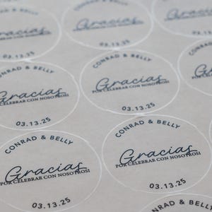 May include: A sheet of circular, semi-transparent stickers featuring black text. Each sticker displays the names Conrad & Belly, the word Gracias, the phrase POR CELEBRAR CON NOSOTROS, and the date 03.13.25 in a clean, modern font layout.