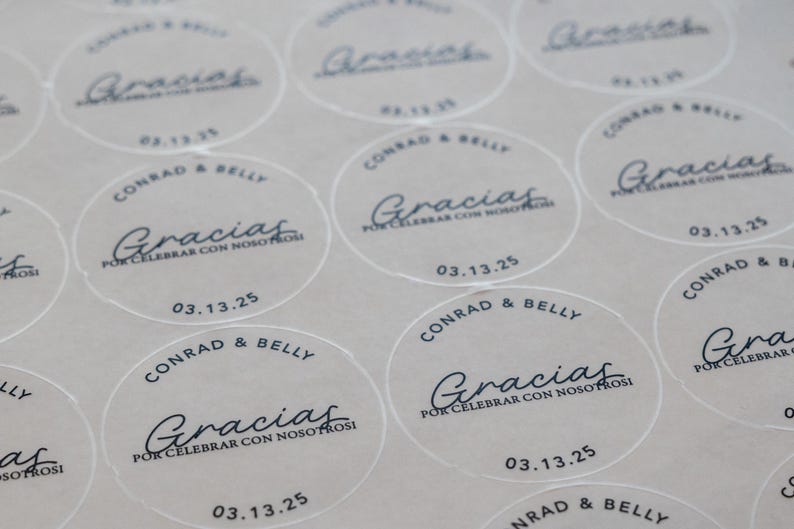 May include: A sheet of circular, semi-transparent stickers featuring black text. Each sticker displays the names Conrad & Belly, the word Gracias, the phrase POR CELEBRAR CON NOSOTROS, and the date 03.13.25 in a clean, modern font layout.