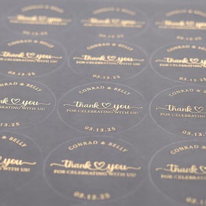 Peut inclure: Gros plan sur une feuille d'autocollants ronds transparents avec du texte dor&eacute;. Chaque autocollant indique "Thank You" avec un c&oelig;ur, les noms "Conrad & Belly" et la date "03.13.25". Pour un mariage ou une c&eacute;l&eacute;bration.