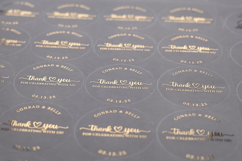 Peut inclure: Gros plan sur une feuille d'autocollants ronds transparents avec du texte dor&eacute;. Chaque autocollant indique "Thank You" avec un c&oelig;ur, les noms "Conrad & Belly" et la date "03.13.25". Pour un mariage ou une c&eacute;l&eacute;bration.