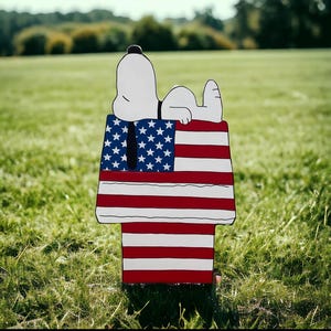 May include: A wooden yard decoration featuring Snoopy lying on top of his doghouse, which is painted with the American flag. The doghouse has red and white stripes and a blue section with white stars. The background is a grassy field.