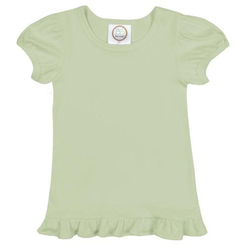 May include: A light green short-sleeved t-shirt with a ruffled hem. The shirt has a round neckline and a small, round label with the text "Baby Bella Boutique" on the front.