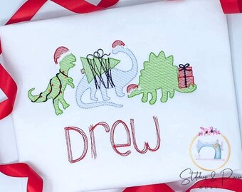 May include: A white embroidered t-shirt with a Christmas theme featuring three dinosaurs wearing Santa hats. The dinosaurs are carrying a Christmas tree and a gift. The name "Drew" is embroidered below the dinosaurs.