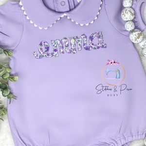 May include: A lavender baby onesie with a white scalloped collar and the name "Emma" embroidered in purple floral fabric. The onesie has a snap closure at the bottom and is decorated with a small sewing machine logo that says "Stitches & Pixie Dust".