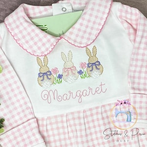 May include: A white baby onesie with pink gingham trim and long sleeves. The onesie has an embroidered design of three bunnies with bows and the name "Margaret" underneath.