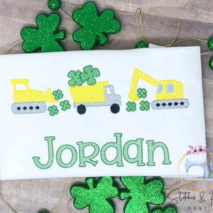 May include: A white t-shirt with an embroidered design of two yellow construction vehicles, a bulldozer and an excavator, with green shamrocks. The name "Jordan" is embroidered below the vehicles in green.