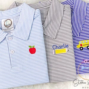 May include: Three striped polo shirts with embroidered designs. The blue shirt has a school bus, the gray shirt has a pencil and the text "Charlie", and the light blue shirt has an apple.