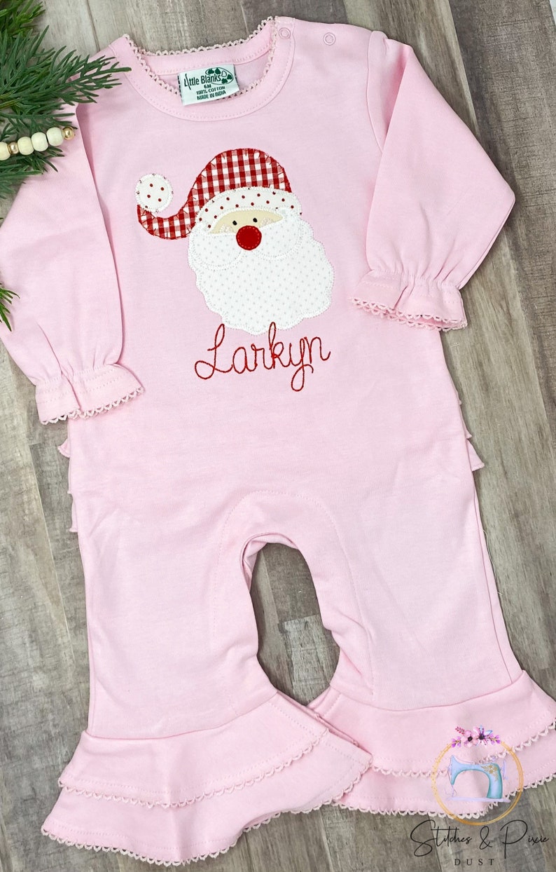 May include: Pink baby onesie with a red and white Santa applique and the name "Larkyn" embroidered in red.