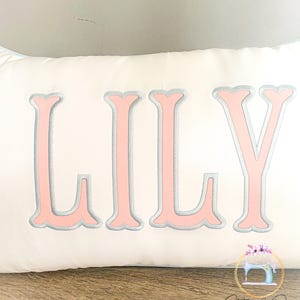 May include: A white rectangular pillow embroidered with the name "LILY" in large, pink, serif letters outlined in gray. The pillow is on a wooden surface, with a logo in the bottom right corner.