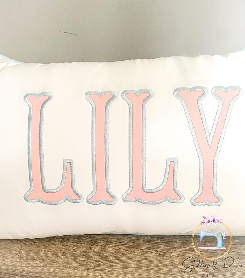 May include: A white rectangular pillow embroidered with the name "LILY" in large, pink, serif letters outlined in gray. The pillow is on a wooden surface, with a logo in the bottom right corner.