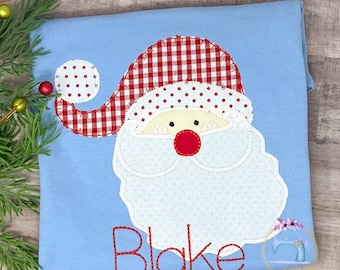 Może przedstawiać: Jasno błękitny t-shirt z aplikacją Świętego Mikołaja w czerwono-białą kratę. Święty Mikołaj ma białą brodę i czerwony nos. Imię "Blake" jest wyszyte na czerwono pod Świętym Mikołajem.