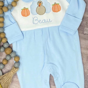 May include: A light blue baby onesie with a white collar and long sleeves. The onesie has an embroidered design of a turkey with pumpkins and the name "Beau".