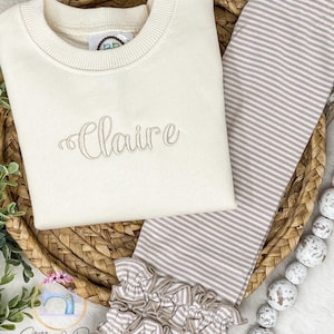 May include: A cream-colored sweatshirt with the name "Claire" embroidered on the front, paired with brown and white striped leggings with a ruffled hem.
