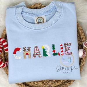 May include: Light blue sweatshirt with the name "CHARLIE" embroidered in colorful festive Christmas themed letters. The letters are decorated with reindeer, candy canes, Christmas trees, and plaid patterns. The sweatshirt also has a small logo that says "Stitches & Pixie Dust".