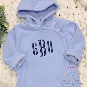 May include: A light blue fleece pullover hoodie featuring a wrap-style neckline with two fabric tabs and long sleeves with folded cuffs. The center of the chest is embroidered with the dark blue monogram letters GBD in a vertical striped font.