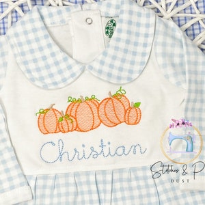 May include: A white long-sleeved baby onesie with a blue and white gingham pattern. The onesie has a Peter Pan collar and features an embroidered design of pumpkins and the name "Christian".