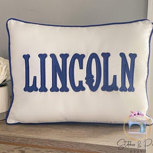 May include: White pillow with a blue embroidered border and the name "LINCOLN" in blue lettering.