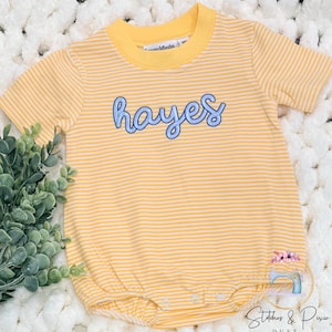 May include: Yellow and white striped baby romper with the name "Hayes" embroidered in blue script. The romper has short sleeves and a round neckline. The bottom has snap closures. The romper is on a white, textured surface.