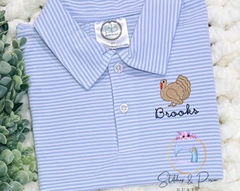 May include: A light blue and white striped polo shirt featuring an embroidered brown turkey and the name Brooks in dark blue script. The shirt has a collar, two white buttons, and a white tag at the neckline labelled BB boutique size 4T.